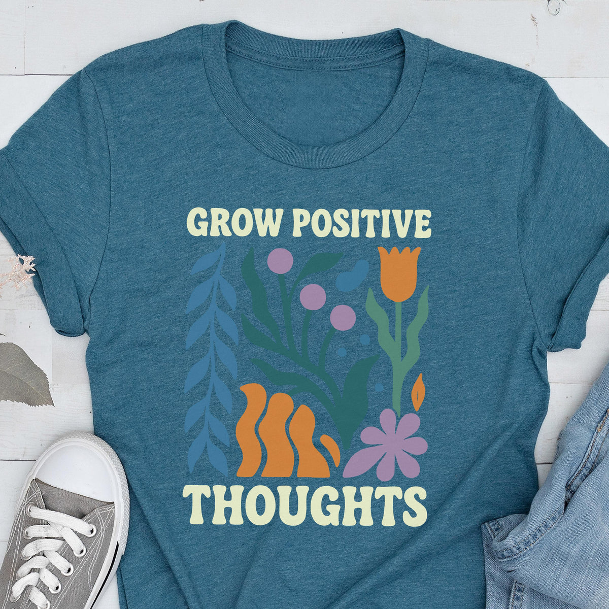May include: Teal t-shirt with the words "GROW POSITIVE THOUGHTS" in a cream color. The shirt features a floral design with orange, purple, and blue elements. A gray sneaker is visible in the lower left corner.