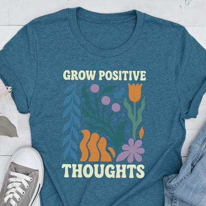 May include: Teal t-shirt with the words "GROW POSITIVE THOUGHTS" in a cream color. The shirt features a floral design with orange, purple, and blue elements. A gray sneaker is visible in the lower left corner.