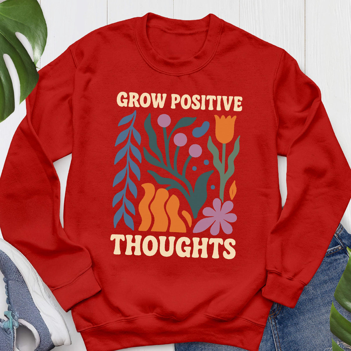 May include: A red sweatshirt with the words "GROW POSITIVE THOUGHTS" in a cream-colored font. The design includes colorful floral and abstract shapes in blue, orange, purple, and pink. The sweatshirt is a crew neck style.