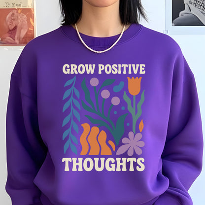 May include: A purple sweatshirt with the words "GROW POSITIVE THOUGHTS" in white. The graphic includes colorful illustrations of flowers and leaves in orange, green, and purple. The sweatshirt has a crew neck.