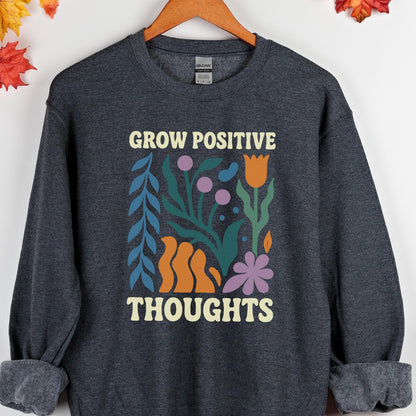 May include: A dark gray sweatshirt with the words "GROW POSITIVE THOUGHTS" in a cream-colored font. The graphic features colorful floral and abstract shapes in blue, orange, purple, and green.