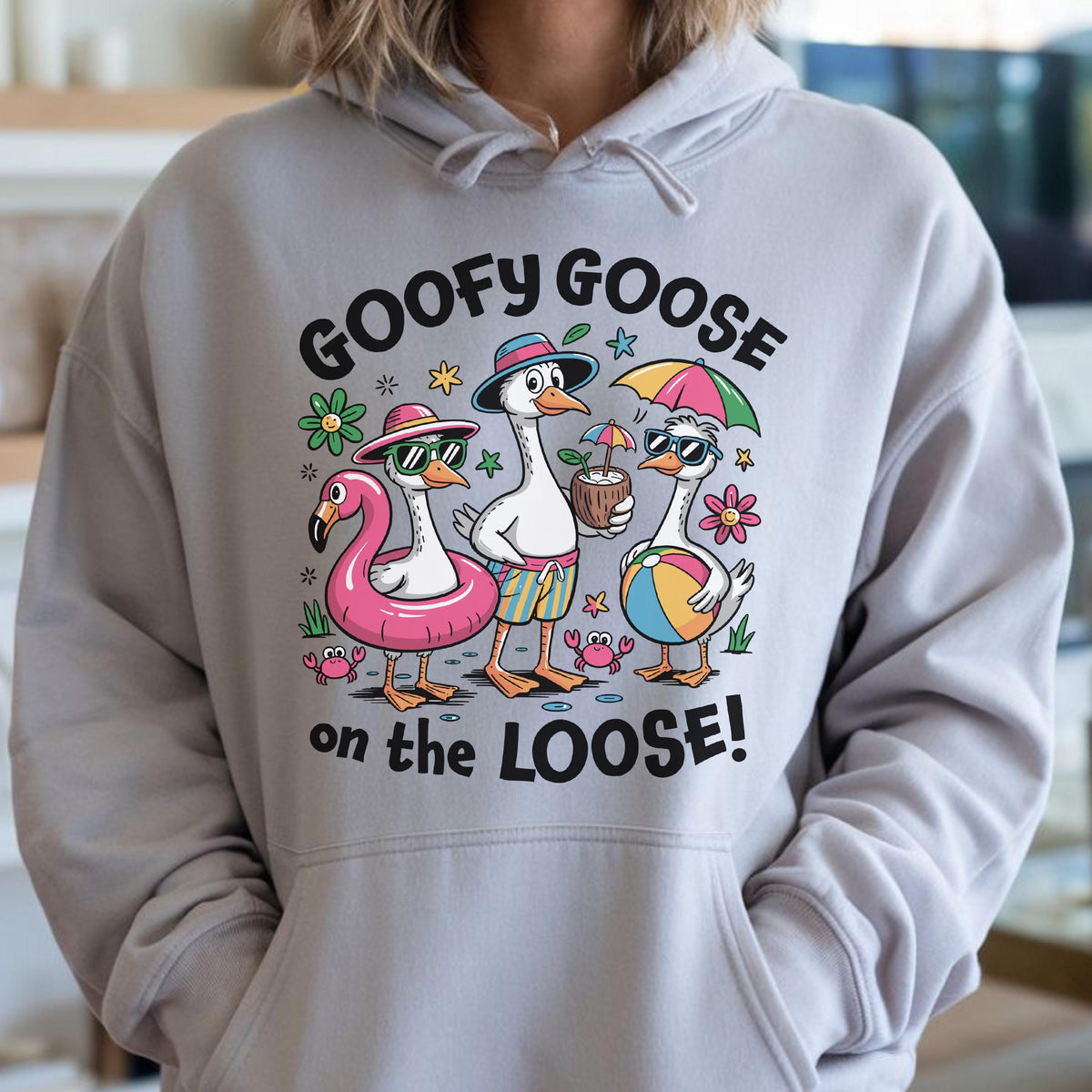 May include: A light gray hoodie featuring a graphic of three cartoon geese in sunglasses and beach attire. The text "GOOFY GOOSE on the LOOSE!" is printed above and below the geese. The geese are wearing a flamingo float, holding a coconut drink, and a beach ball.