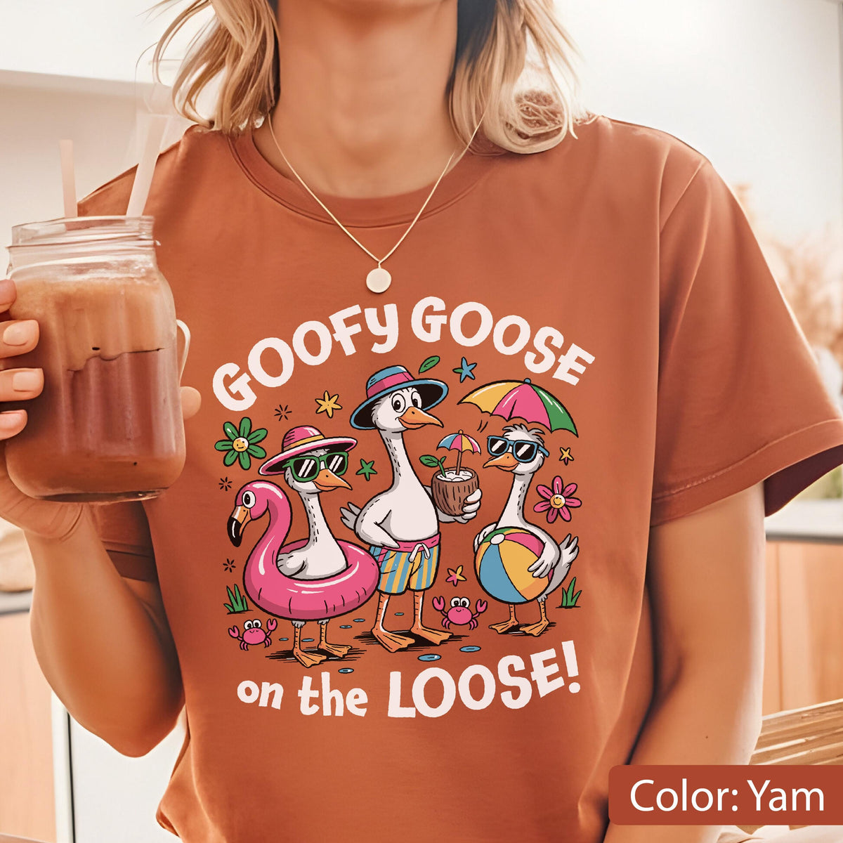 May include: A yam-colored t-shirt featuring a graphic of three geese on vacation. The text reads GOOFY GOOSE on the LOOSE! The geese wear hats and sunglasses, with one in a pink flamingo float, one holding a coconut drink, and one holding a beach ball.