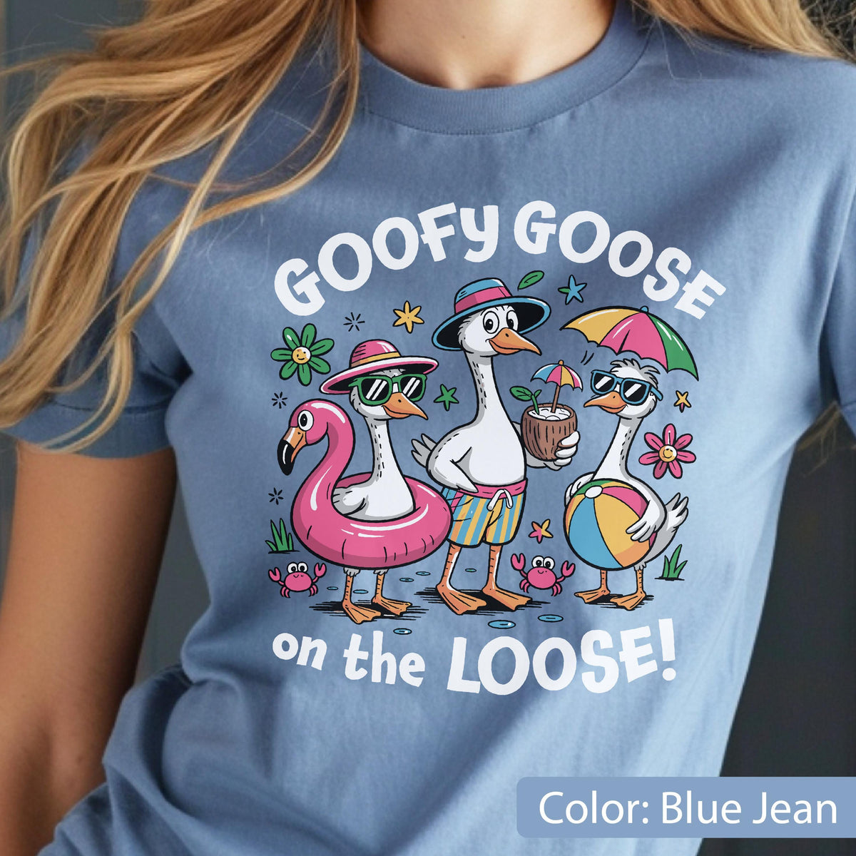 May include: A blue jean colored t-shirt featuring a graphic of three geese on vacation. The text reads GOOFY GOOSE on the LOOSE! The geese are wearing sunglasses, hats, and swim trunks, with a pink flamingo float, a coconut drink, and a beach ball.