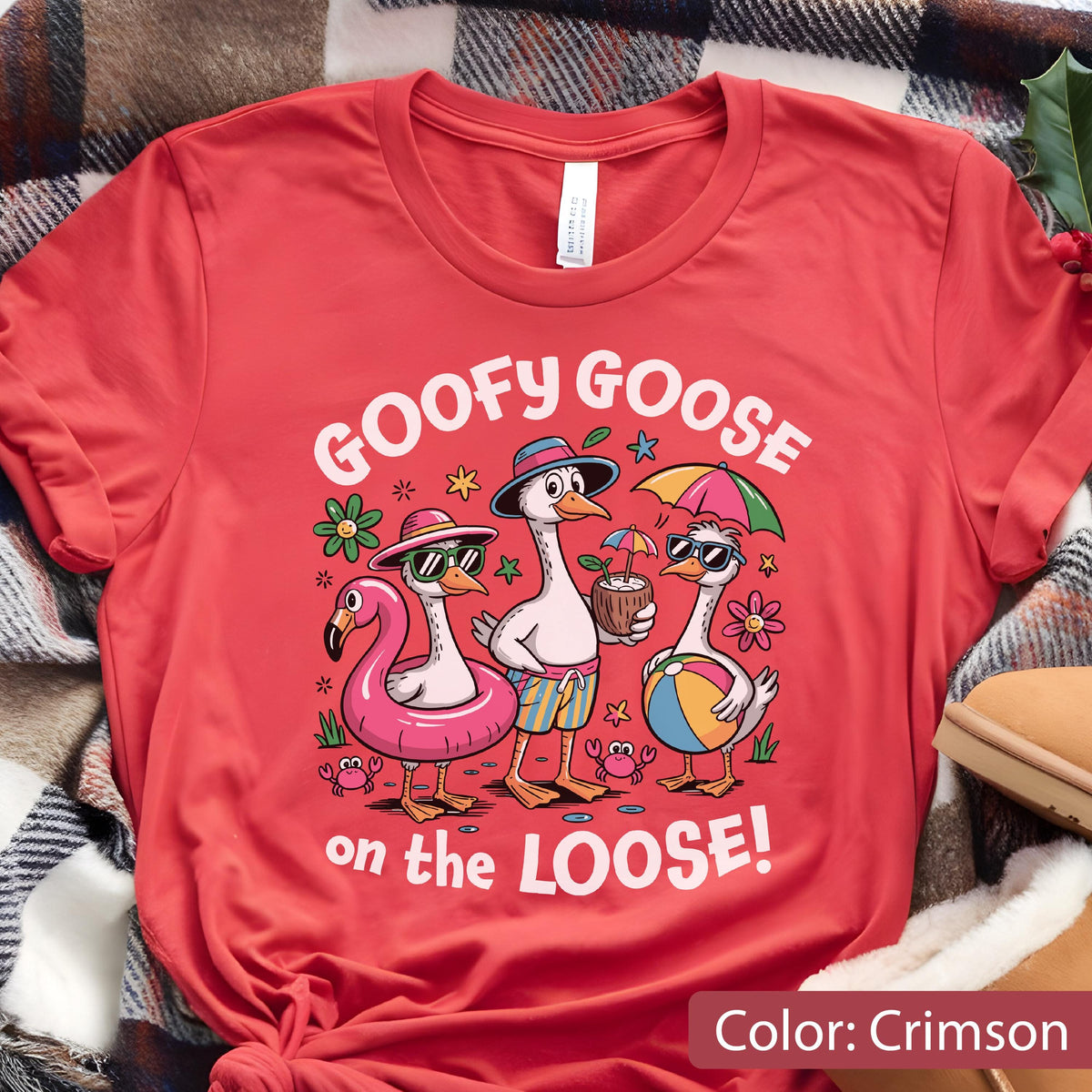 May include: A crimson t-shirt featuring a graphic of three geese on a beach. The geese wear sunglasses and hats, with one in a pink floatie, one holding a coconut drink, and one holding a beach ball. The text reads GOOFY GOOSE on the LOOSE!