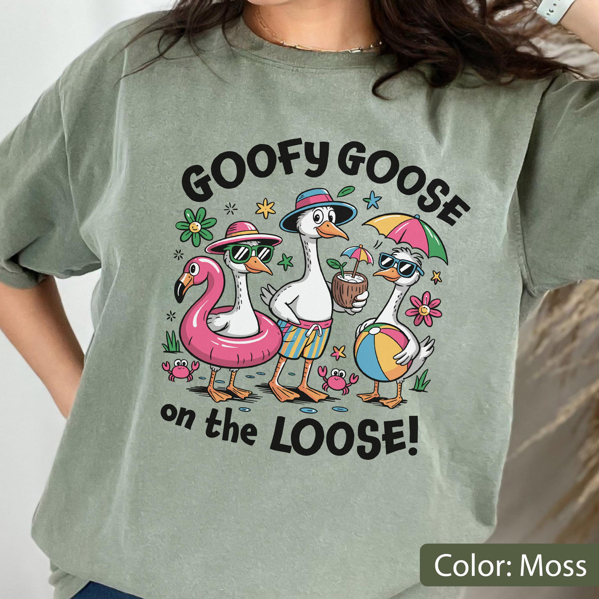 May include: A moss green t-shirt featuring a graphic of three geese on vacation. The text reads GOOFY GOOSE on the LOOSE! The geese wear sunglasses, hats, and swim trunks, while holding a pink flamingo float, a coconut drink, and a beach ball.