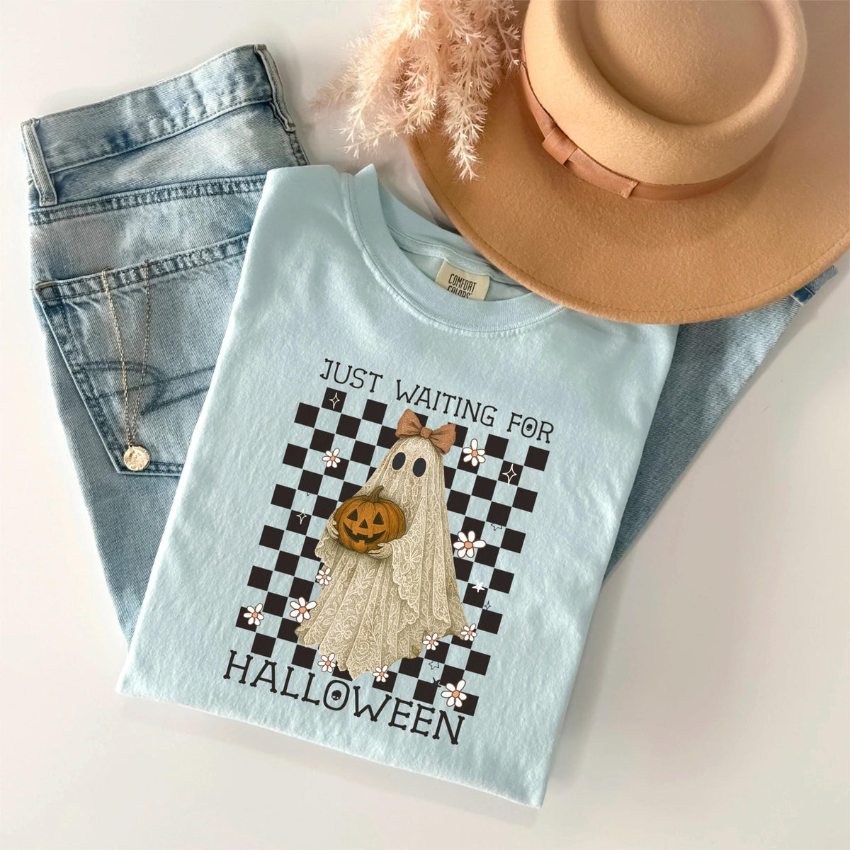 May include: A light blue t-shirt with the text "JUST WAITING FOR HALLOWEEN" and a graphic of a ghost holding a pumpkin. The shirt is paired with light wash jeans, a tan hat, and a gold necklace.
