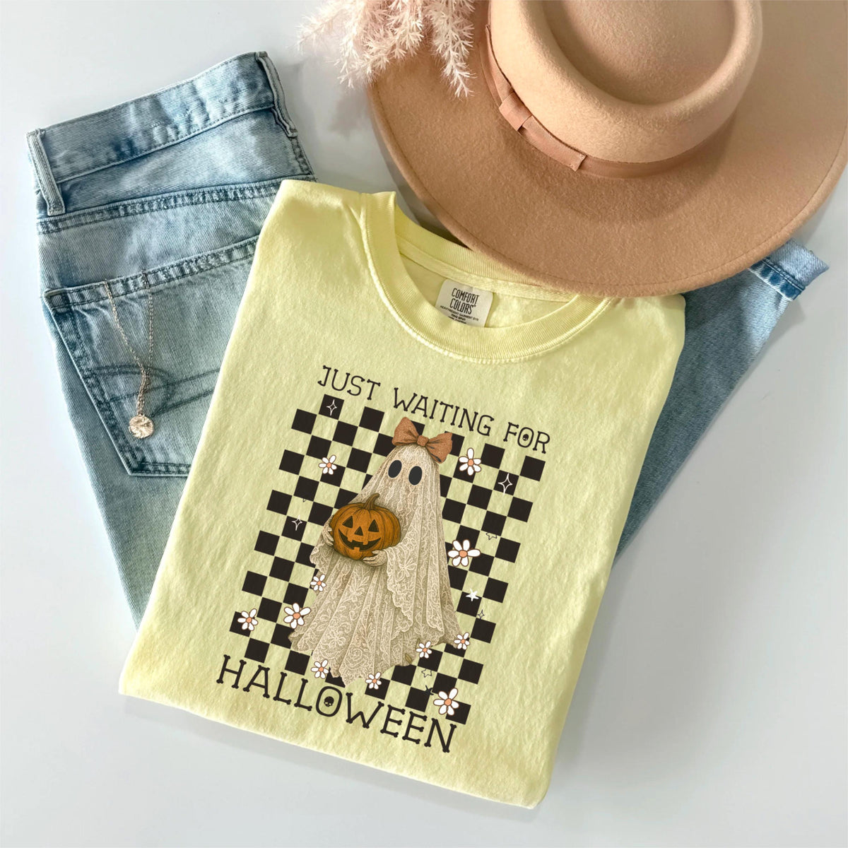 May include: A light yellow t-shirt with a Halloween ghost graphic, text that reads "JUST WAITING FOR HALLOWEEN", and a brown hat and blue jeans. The ghost graphic includes a pumpkin and bow. The shirt is folded on top of the jeans.