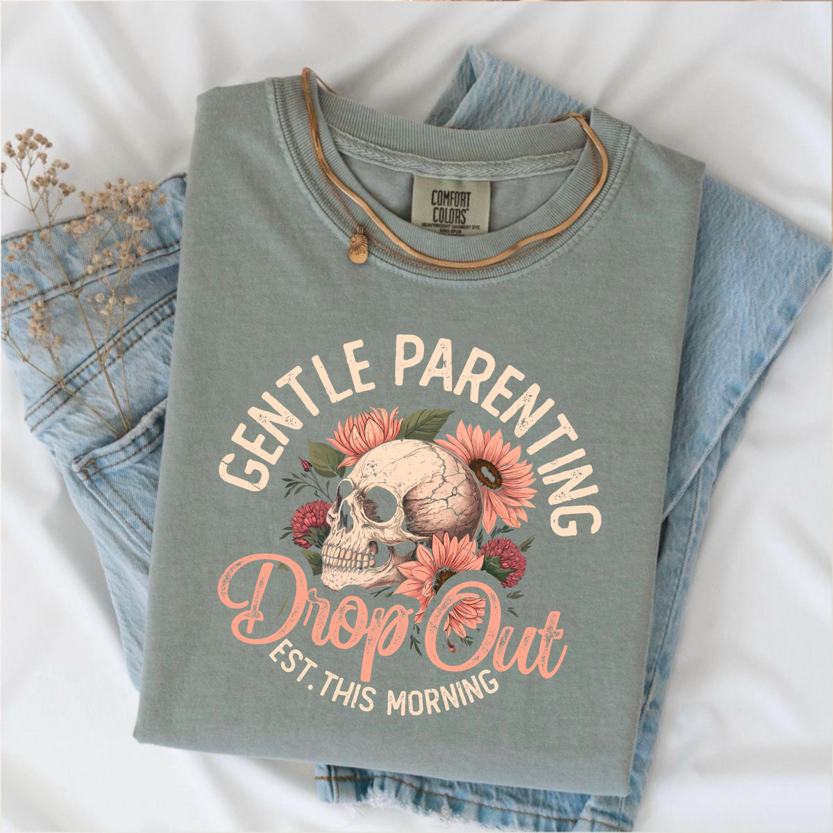 May include: Sage green t-shirt with the words "GENTLE PARENTING" and "Drop Out" in a floral design with a skull. The shirt is folded on top of blue jeans and a gold necklace.
