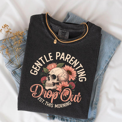 May include: Dark gray t-shirt with the words "GENTLE PARENTING" arched above a skull and floral design. Below the skull, the text reads "Drop Out Est. This Morning". A gold necklace rests on the shirt, which is folded on top of blue jeans.