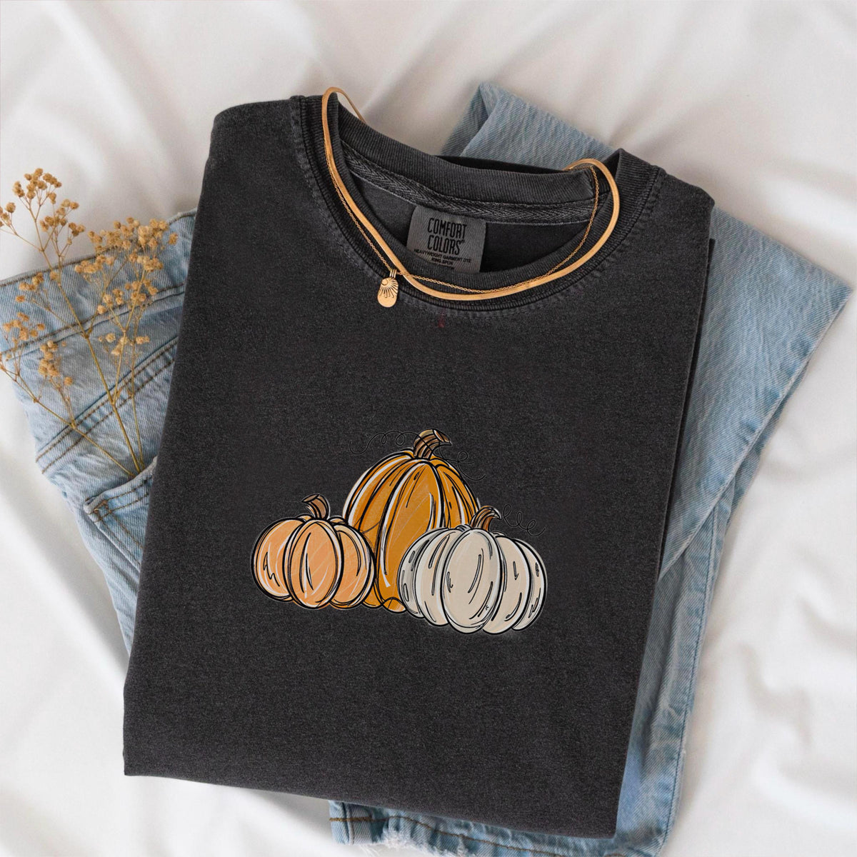 May include: A dark gray t-shirt with a pumpkin graphic. The shirt features three pumpkins in shades of orange and white. A gold necklace with a small pendant rests on the shirt. The shirt is folded on top of a pair of blue jeans.