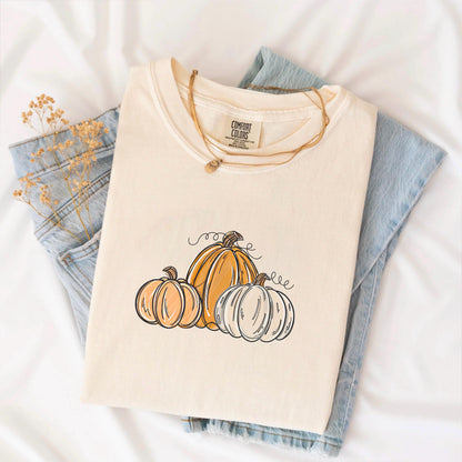 May include: Cream-colored t-shirt featuring a pumpkin graphic. The design includes three pumpkins in shades of orange and white with black outlines and curly vines. The shirt is folded on top of blue jeans.