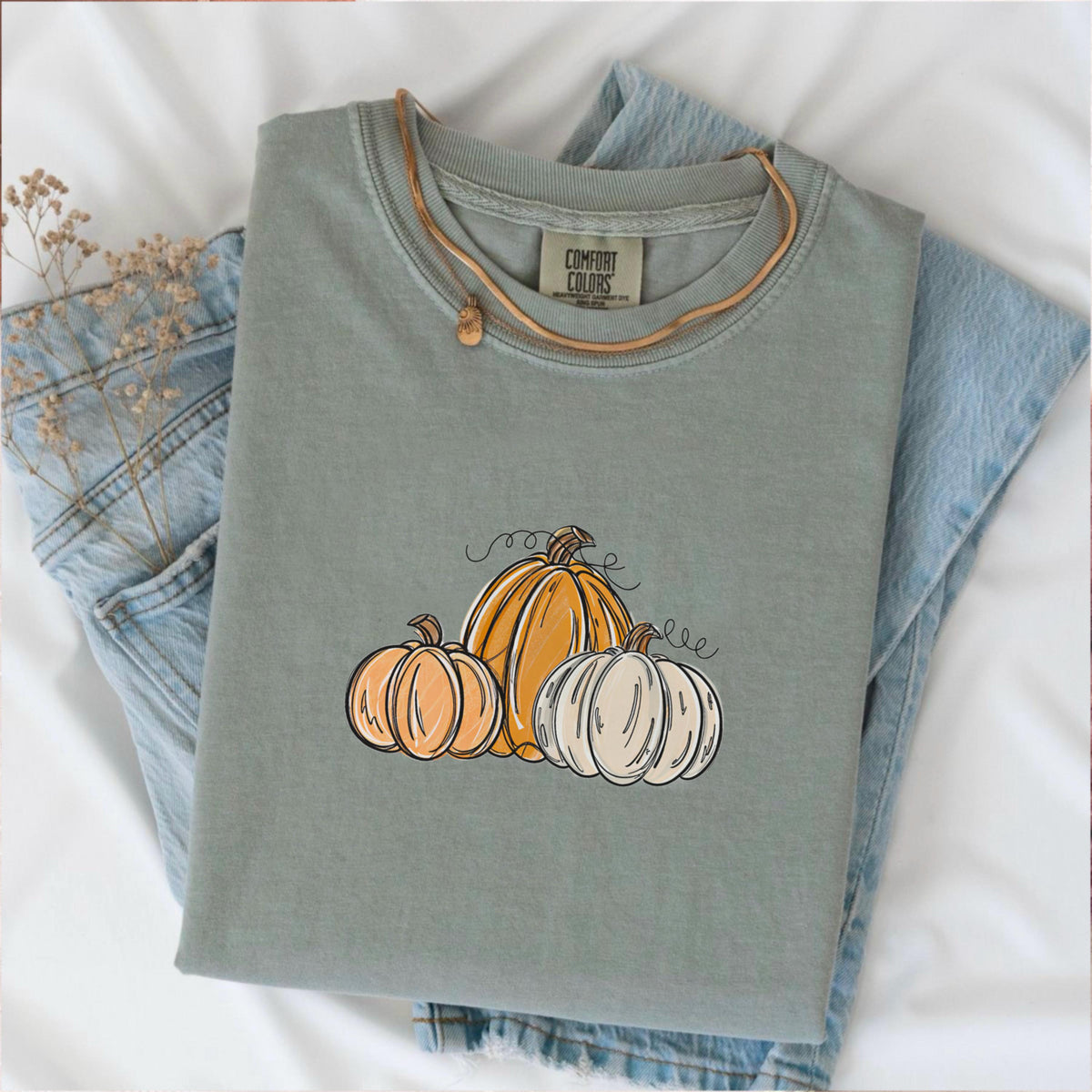 May include: Sage green Comfort Colors t-shirt with a pumpkin graphic. The graphic features three pumpkins in shades of orange and white with black outlines and curly vines. The shirt is folded on top of a pair of blue jeans.