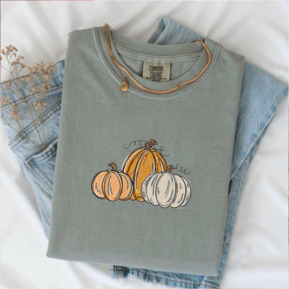 May include: Sage green Comfort Colors t-shirt with a pumpkin graphic. The graphic features three pumpkins in shades of orange and white with black outlines and curly vines. The shirt is folded on top of a pair of blue jeans.