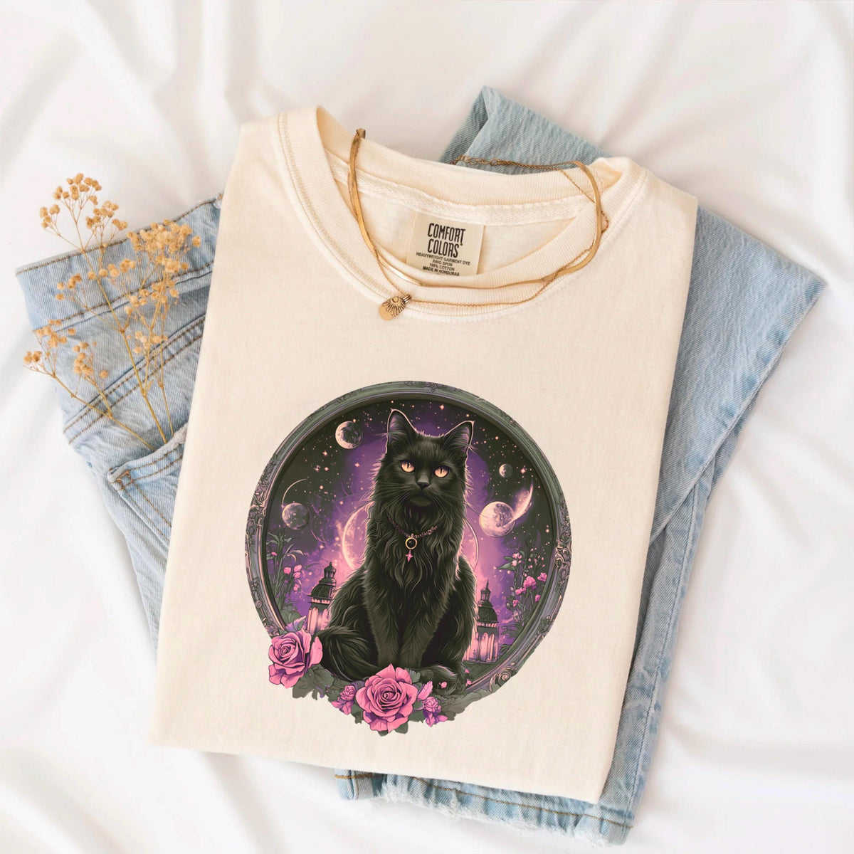 May include: Cream-colored t-shirt featuring a black cat illustration within a circular frame. The cat has orange eyes and is surrounded by a purple and pink celestial scene with roses. The shirt is folded on top of blue jeans.