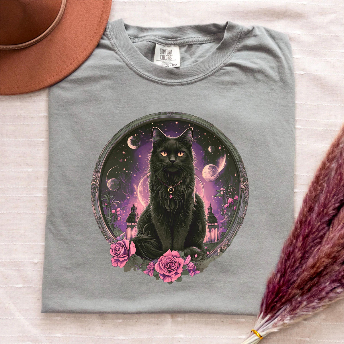 May include: A gray Comfort Colors t-shirt featuring a circular graphic of a black cat with orange eyes, a necklace, and a celestial background with moons and planets. Pink roses surround the cat. A brown hat and purple dried flowers are visible.