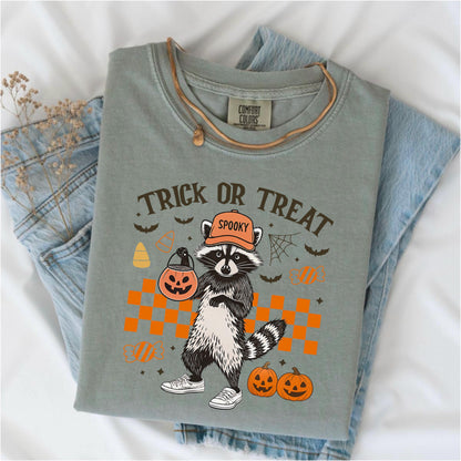 May include: Sage green t-shirt with a Halloween-themed graphic. The design features a raccoon wearing an orange hat and holding a jack-o'-lantern. The text "Trick or Treat" is above the raccoon, with bats, candy corn, and pumpkins.