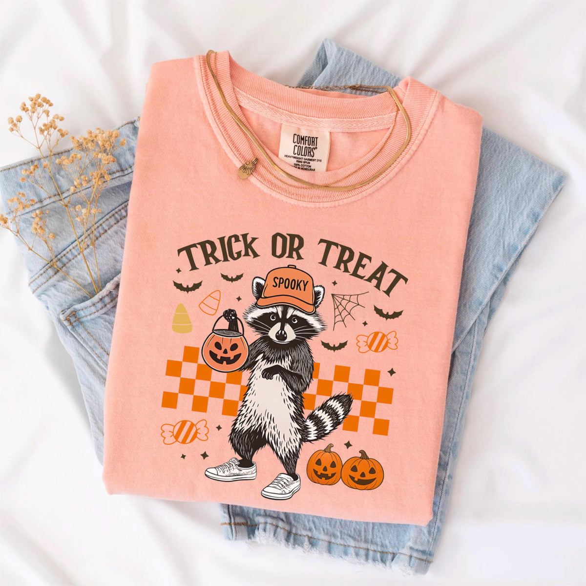 May include: A peach-colored t-shirt with a Halloween-themed graphic. The design features a raccoon wearing a hat and sneakers, holding a jack-o'-lantern bucket. The text "Trick or Treat" and "Spooky" are also displayed. The shirt is folded on top of blue jeans.