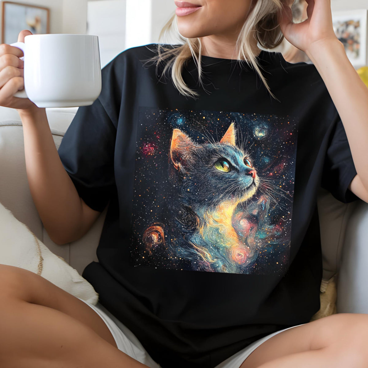 May include: Black t-shirt featuring a vibrant, artistic depiction of a cat's face set against a cosmic background of stars and galaxies. The cat has yellow eyes and orange-tipped ears. The shirt is worn by a person holding a white mug.