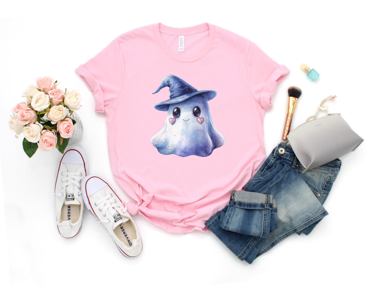 May include: A light pink t-shirt featuring a watercolor illustration of a friendly ghost wearing a blue witch hat. The ghost has heart-shaped cheeks. The shirt is styled with blue jeans, white sneakers, and makeup.