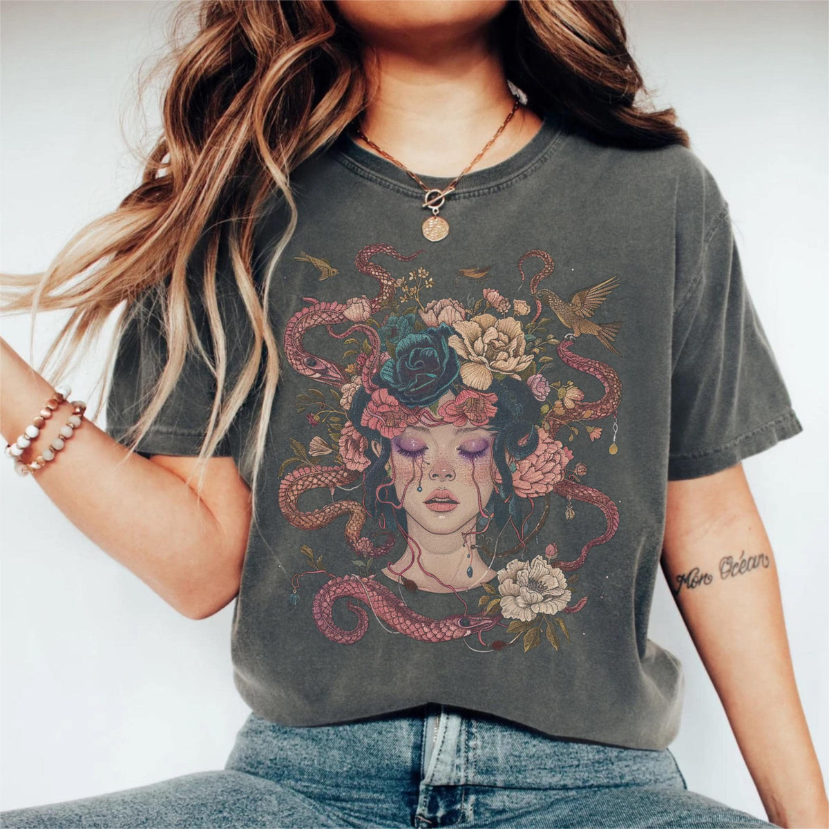 May include: Dark gray t-shirt featuring a detailed illustration of a woman's face with closed eyes, adorned with flowers, snakes, and birds. The artwork includes pink, blue, and beige tones, creating a unique and artistic design. The shirt is a comfortable fit.