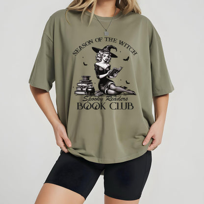May include: Olive green t-shirt with a graphic featuring a witch reading a book, with the text "SEASON OF THE WITCH" and "Spooky Readers BOOK CLUB". The witch is wearing a hat and sitting near a stack of books.