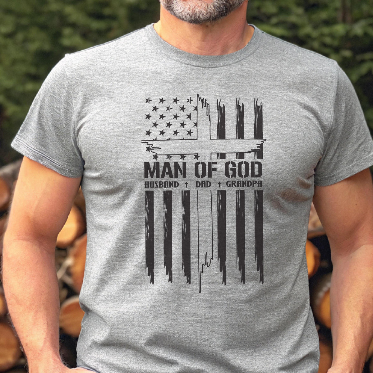 May include: A gray t-shirt featuring a distressed American flag design with the text "MAN OF GOD" above the words "HUSBAND + DAD + GRANDPA" in black. The shirt has short sleeves and a crew neck.
