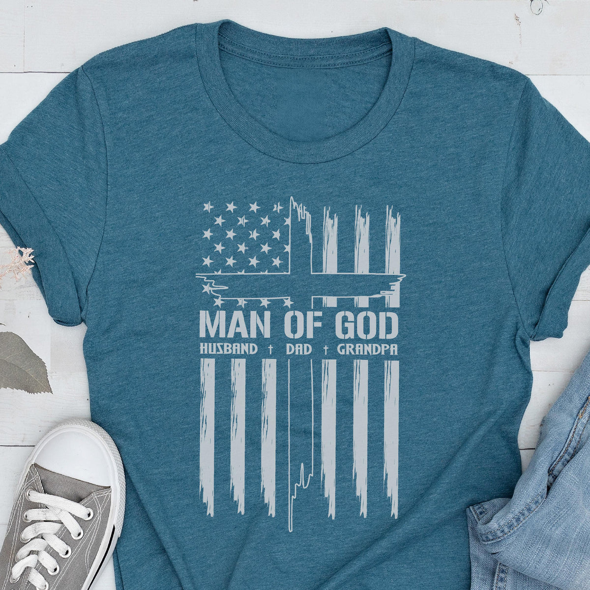 May include: Teal t-shirt featuring a distressed white graphic of the American flag with a cross and the words "MAN OF GOD, HUSBAND, DAD, GRANDPA." The shirt is made of a soft material and has short sleeves.