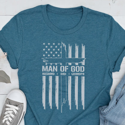 May include: Teal t-shirt featuring a distressed white graphic of the American flag with a cross and the words "MAN OF GOD, HUSBAND, DAD, GRANDPA." The shirt is made of a soft material and has short sleeves.