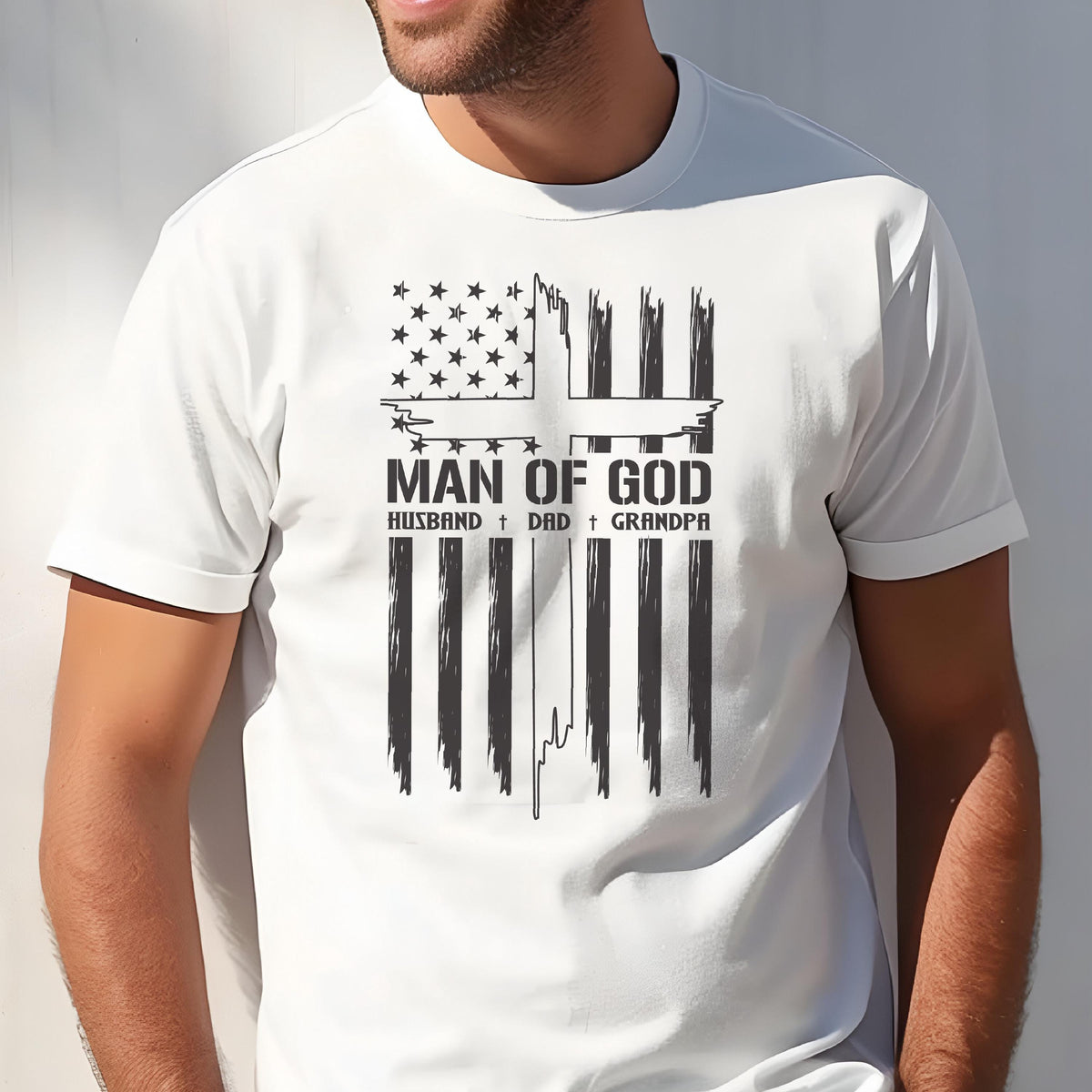 May include: White t-shirt featuring a black and white graphic design. The design includes an American flag motif with a cross, the words "MAN OF GOD", and the words "HUSBAND + DAD + GRANDPA".