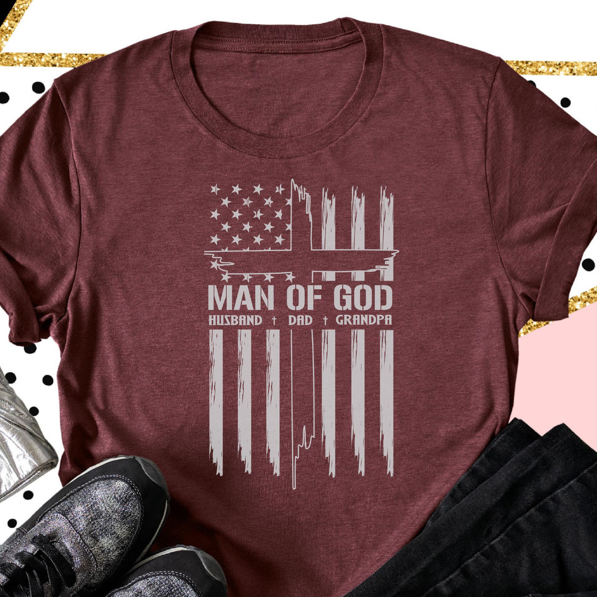 May include: Burgundy t-shirt featuring a distressed American flag design with a cross and the words "MAN OF GOD." Below the text are the words "HUSBAND + DAD + GRANDPA" in a white, distressed font. The shirt is folded.