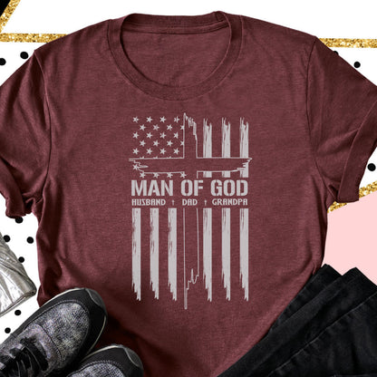 May include: Burgundy t-shirt featuring a distressed American flag design with a cross and the words "MAN OF GOD." Below the text are the words "HUSBAND + DAD + GRANDPA" in a white, distressed font. The shirt is folded.