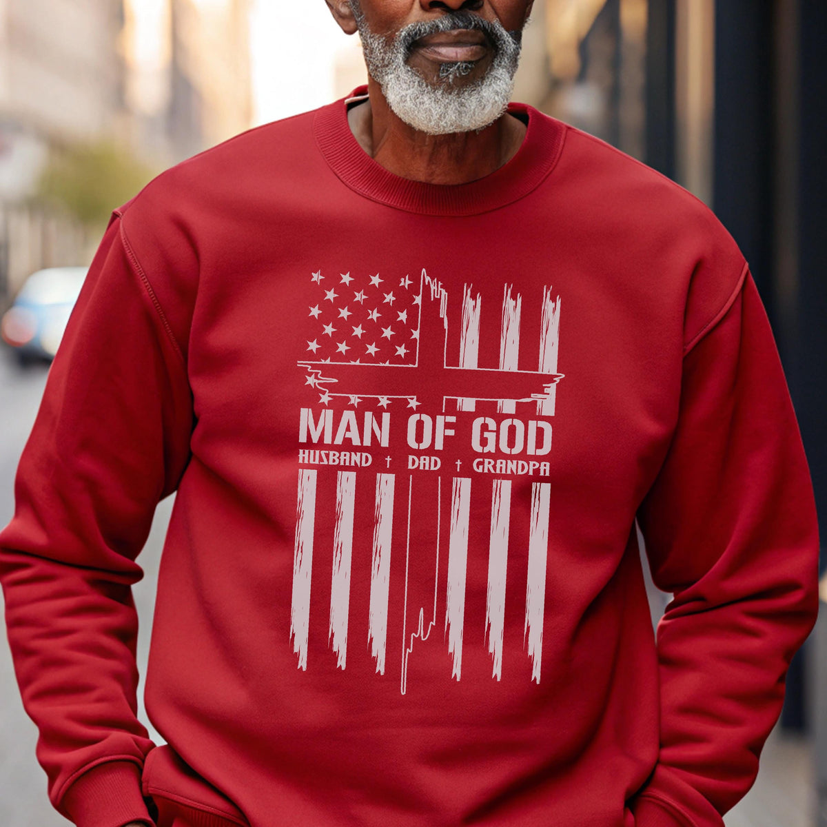 May include: A red sweatshirt with a white graphic design. The design features an American flag with a cross, the words "MAN OF GOD," and the words "HUSBAND + DAD + GRANDPA."