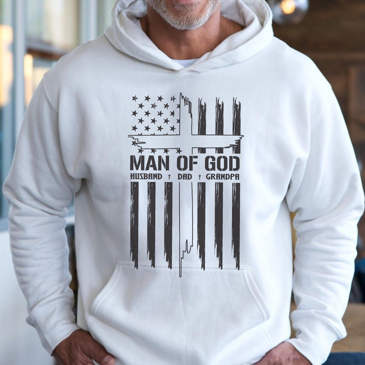 May include: White hooded sweatshirt with a graphic design. The design features an American flag with a cross and the words "MAN OF GOD, HUSBAND, DAD, GRANDPA" in black. The sweatshirt has a front pocket and a drawstring hood.
