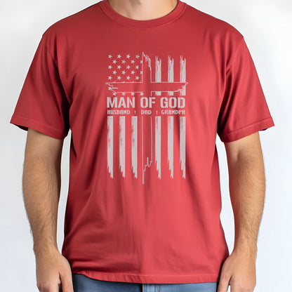 May include: Red t-shirt featuring a distressed American flag design with the text "MAN OF GOD" and the words "HUSBAND + DAD + GRANDPA" in a white, vintage font. The shirt is short-sleeved and has a crew neck.