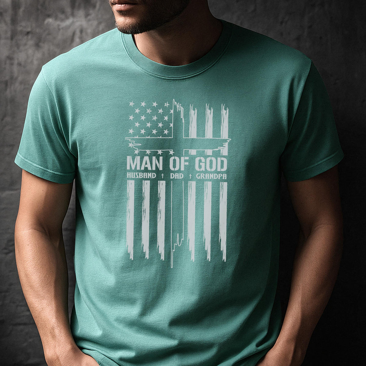 May include: A teal t-shirt with a distressed white graphic of an American flag design. The design includes the text "MAN OF GOD" above "HUSBAND + DAD + GRANDPA". The shirt has short sleeves and a crew neck.