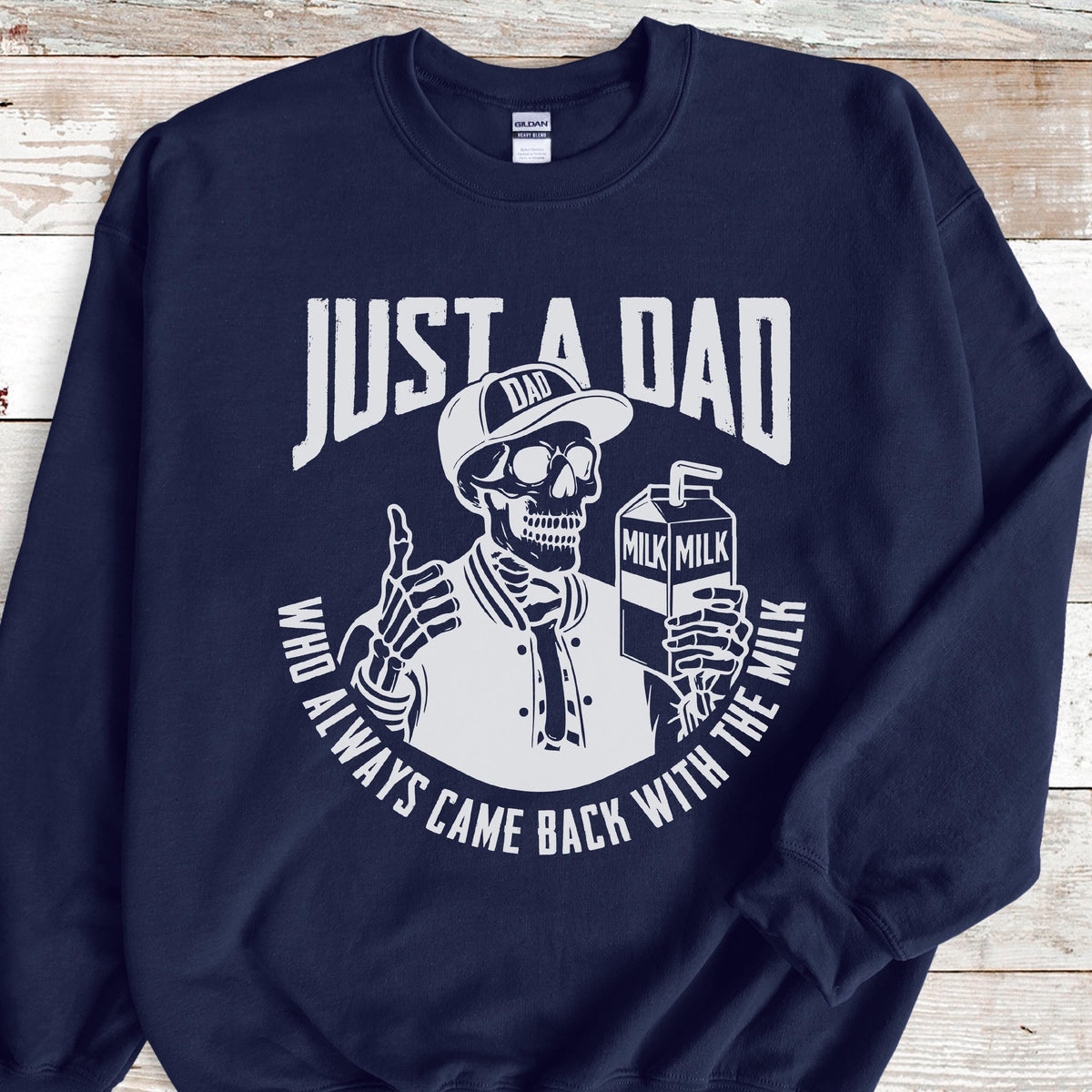 May include: Navy blue sweatshirt with a white graphic. The graphic features a skeleton wearing a baseball cap and jacket, holding a milk carton. Text reads "JUST A DAD" and "WHO ALWAYS CAME BACK WITH THE MILK."