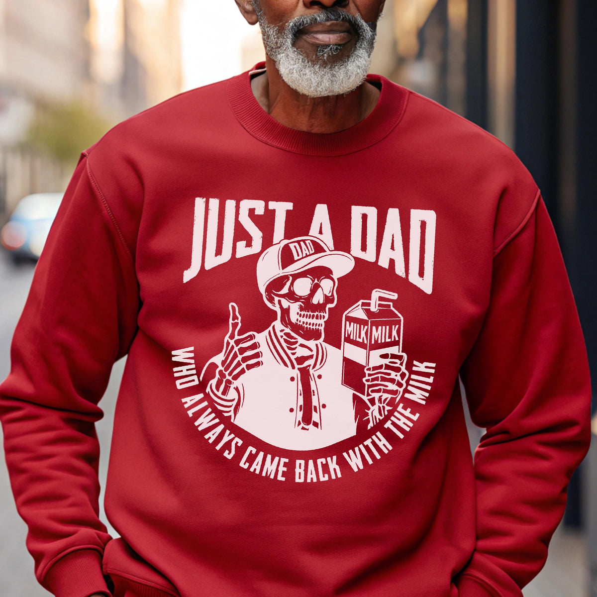 May include: A red sweatshirt with a white graphic that reads "JUST A DAD" above a skeleton wearing a baseball cap and holding a milk carton. The text "WHO ALWAYS CAME BACK WITH THE MILK" is printed around the skeleton.