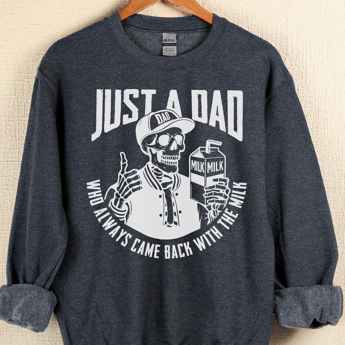 May include: Dark gray sweatshirt with a white graphic of a skeleton wearing a baseball cap and jacket, holding a milk carton. The text reads "JUST A DAD" above the skeleton and "WHO ALWAYS CAME BACK WITH THE MILK" below.