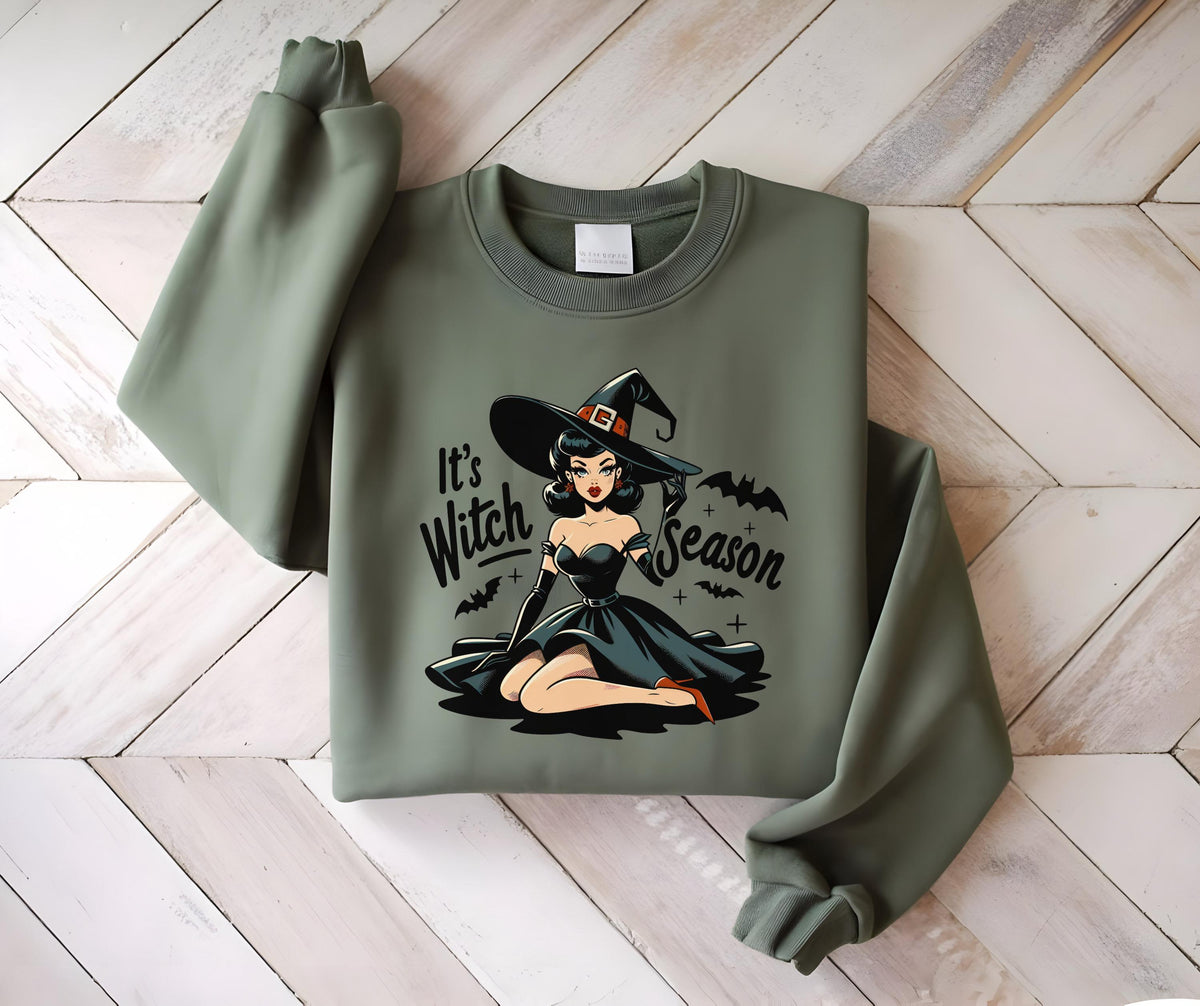 May include: Sage green sweatshirt featuring a retro-style illustration of a witch in a black dress and hat. The text "It's Witch Season" is printed on the front. The design includes bats and crosses, perfect for Halloween.