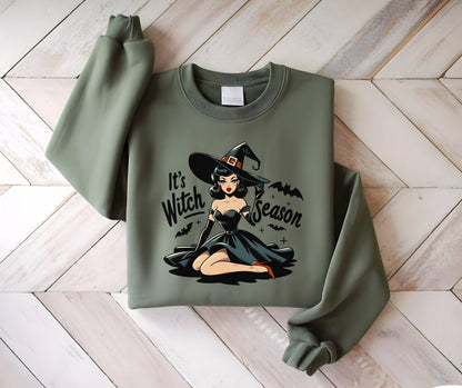 May include: Sage green sweatshirt featuring a retro-style illustration of a witch in a black dress and hat. The text "It's Witch Season" is printed on the front. The design includes bats and crosses, perfect for Halloween.