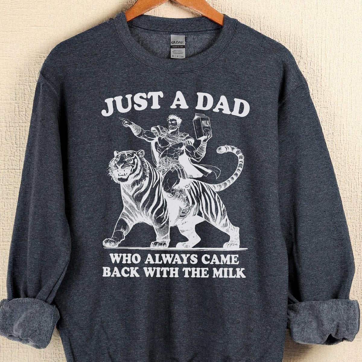 May include: Dark gray sweatshirt with the text "JUST A DAD WHO ALWAYS CAME BACK WITH THE MILK." A white graphic depicts a man riding a tiger, holding a milk carton. The sweatshirt has a crew neck and long sleeves.