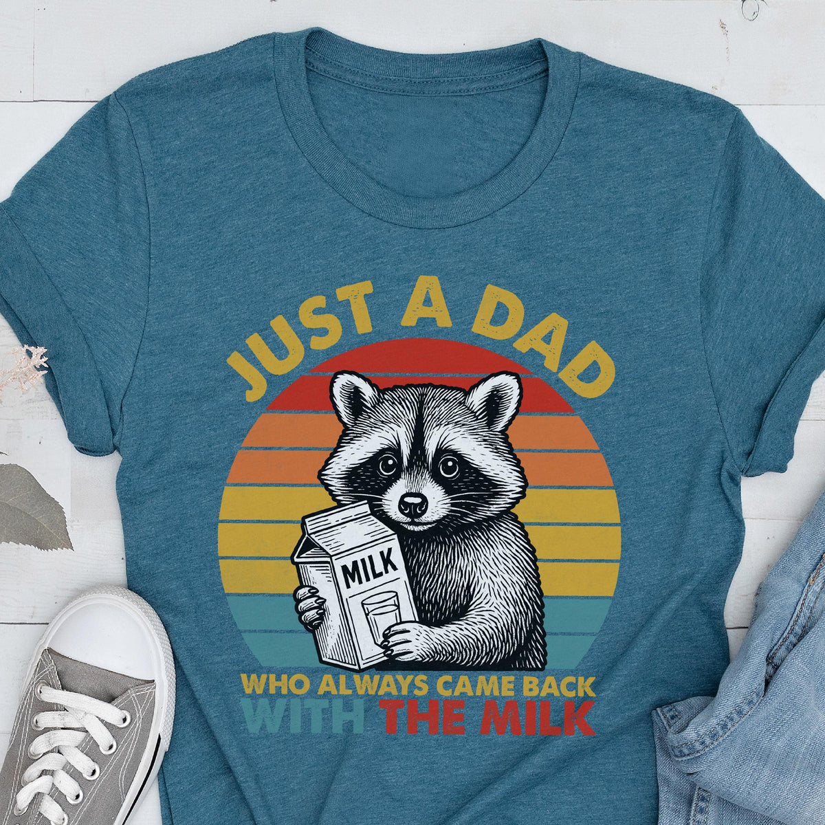 May include: Teal t-shirt with a graphic of a raccoon holding a milk carton. The text reads "JUST A DAD WHO ALWAYS CAME BACK WITH THE MILK" in yellow and red. The raccoon is set against a sunset background.