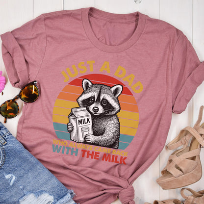 May include: Dusty rose t-shirt featuring a raccoon holding a milk carton and a glass of milk. The text reads "JUST A DAD WHO ALWAYS CAME BACK WITH THE MILK" in yellow and white. The design includes a retro sunset graphic.