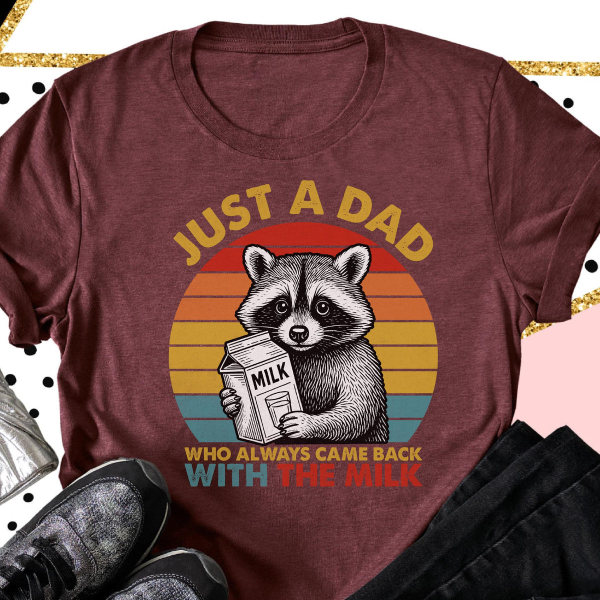 May include: Burgundy t-shirt with a raccoon graphic holding a milk carton. The text reads "JUST A DAD WHO ALWAYS CAME BACK WITH THE MILK" in a retro sunset design. The shirt is a crew neck.