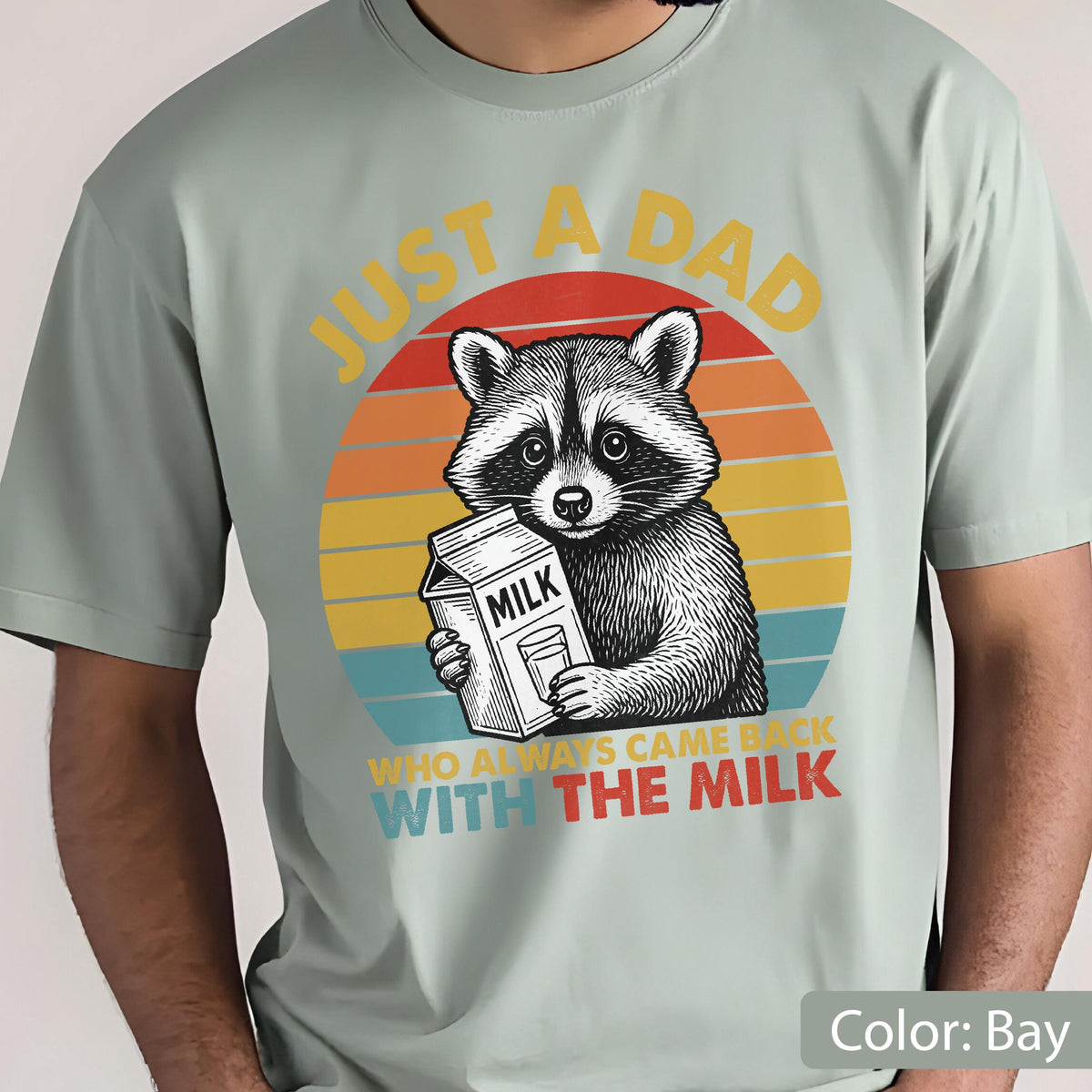 May include: A light green t-shirt featuring a raccoon holding a milk carton. The raccoon is set against a retro sunset design with the text "JUST A DAD WHO ALWAYS CAME BACK WITH THE MILK".