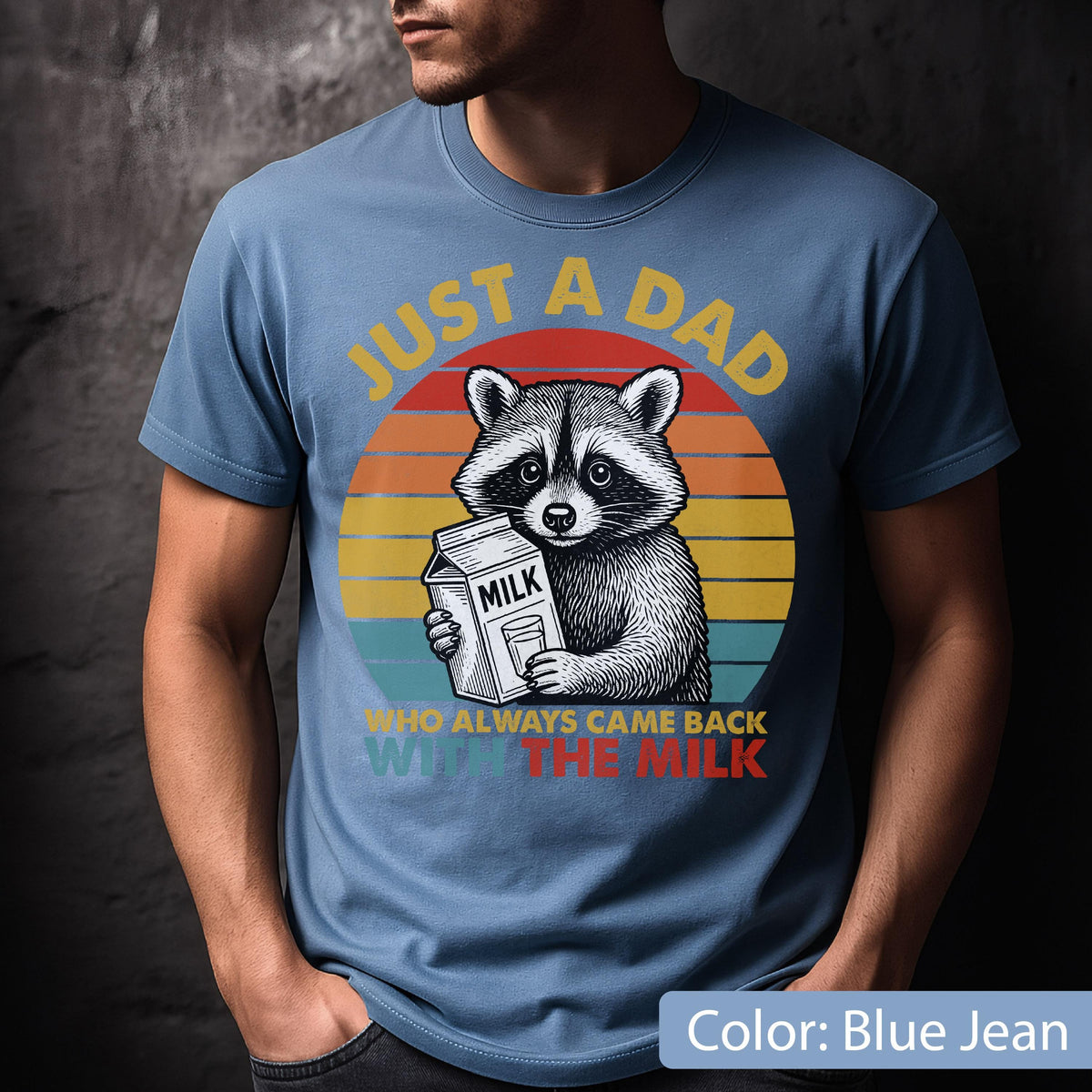 May include: Blue jean t-shirt featuring a raccoon holding a milk carton, set against a sunset design. Text reads "JUST A DAD WHO ALWAYS CAME BACK WITH THE MILK". The graphic is in yellow, red, and orange.
