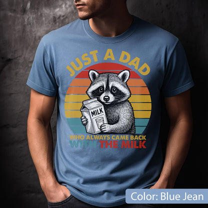 May include: Blue jean t-shirt featuring a raccoon holding a milk carton, set against a sunset design. Text reads "JUST A DAD WHO ALWAYS CAME BACK WITH THE MILK". The graphic is in yellow, red, and orange.