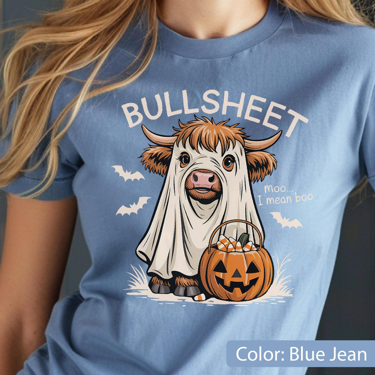 May include: A blue jean colored t-shirt featuring a graphic of a Highland cow wearing a white ghost costume. The text reads BULLSHEET and moo... I mean boo. The cow holds an orange jack-o'-lantern bucket filled with candy corn, surrounded by bats.