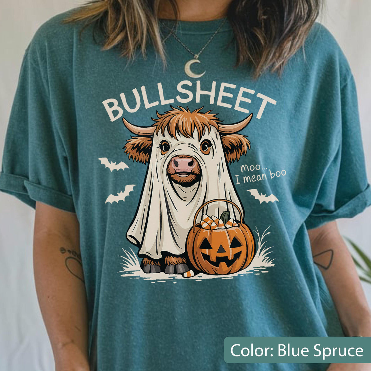 May include: A blue spruce t-shirt featuring a graphic of a Highland cow wearing a white ghost sheet. The text reads BULLSHEET and moo... I mean boo. The cow holds an orange jack-o'-lantern bucket filled with candy corn, surrounded by white bats.